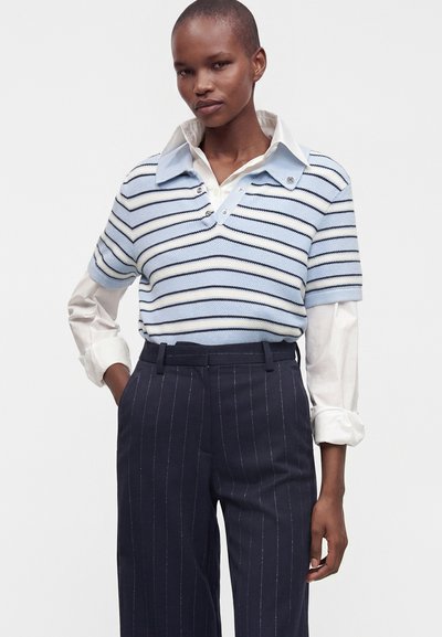 Personne portant un pull à manches courtes rayé bleu clair et blanc par-dessus une chemise blanche à boutons et un pantalon bleu marine à fines rayures.