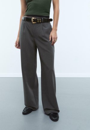 Person trägt weite graue Hosen, schwarzen Ledergürtel mit goldener Schnalle, dunkelgrünes geripptes Tanktop, schwarze Schuhe und goldene Ringe an den Fingern.