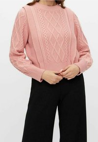 Femme portant un pull rose à torsades à manches longues et un pantalon noir à jambes larges, les mains jointes à la taille.