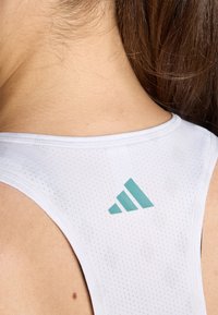 Bjela sportska majica bez rukava s prozračnom mrežastom teksturom, s svijetloplavim Adidas logotipom na leđima blizu izreza.