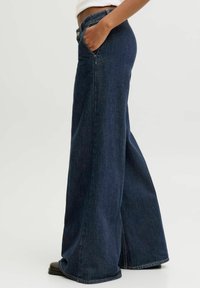 Person wearing dark blue wide-leg jeans with one hand in pocket, paired with black shoes and a white cropped top.