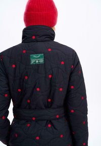 Prešívaný čierny kabát s motívmi červených jabĺk, s opaskom v páse a zelenou nášivkou na chrbte, štruktúrovaná tkanina a stojatý golier.