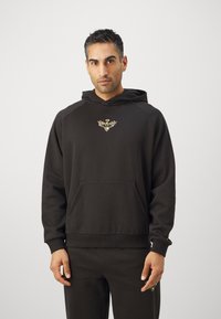 Hanorac negru dintr-un material moale, dotat cu o buzunar frontal și un logo cu detalii intricate pe piept. Decolteu rotund și croială lejeră.