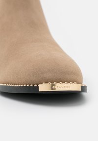 Botín de ante marrón claro con punta redondeada, herrajes en tono dorado, detalles de cuentas a lo largo de la suela y nombre de la marca en relieve.