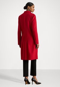 Palton roșu din lână, cu croi drept, mâneci lungi și guler cu decupaj. Asortat cu pantaloni negri scurți și pantofi cu toc înalt.