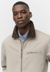 Veste beige avec un col en velours côtelé marron et une poche zippée. Porte sur une chemise rayée bleu clair et blanche. Texture de tissu lisse.