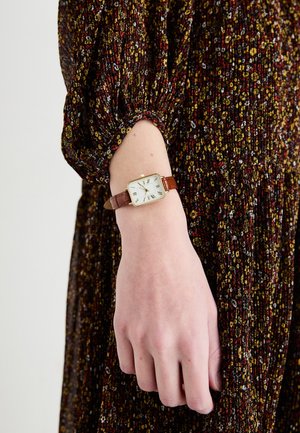 Poignet avec montre carrée en or et bracelet en cuir marron sur une personne portant une robe à manches longues à motif floral.