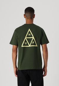 T-shirt en coton vert kaki avec un grand motif en triangle beige dans le dos, contenant les lettres H, U et F disposées à l'intérieur du triangle.