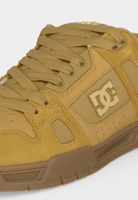 Gros plan d'une basket DC beige avec des lacets épais, des panneaux en suède et en mesh, et une semelle en caoutchouc. Le logo DC est visible sur le côté.