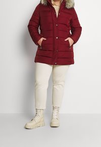 Röd puffjacka med huva av fuskpäls, dragkedjefickor och tryckknappsstängning; bärs med krämfärgade byxor och beige snörstövlar med kraftig sula.