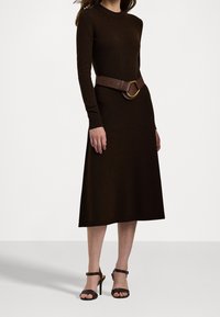 Brun stickad klänning med långa ärmar och kroppsnära liv, prydd med ett brett brunt bälte med en guldfinis. Klänningen fulländas av högklackade sandaler.