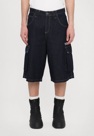 Shorts cargo en denim bleu foncé avec poches latérales, couture contrastante et coupe droite. Associés à des bottes montantes noires et des chaussettes blanches.