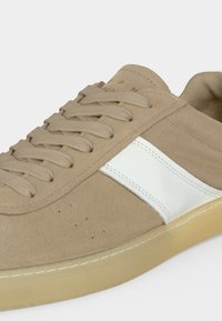 Baskets en daim beige avec lacets beiges, bande latérale en cuir blanc et semelle en caoutchouc beige semi-translucide sur fond blanc.
