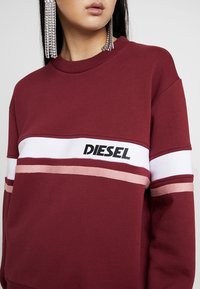 Felpa color bordeaux con una striscia orizzontale bianca e dettagli rosa, impreziosita dal logo "DIESEL" in nero. Materiale morbido in misto cotone.