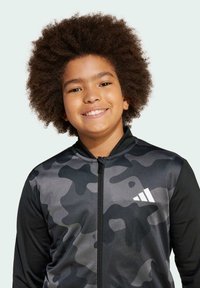 Černá a šedá maskáčová zipová bunda s hladkým povrchem a bílým logem. Design zahrnuje vysoký límec a dlouhé rukávy.