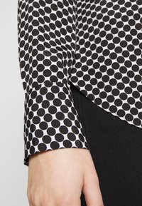 Gros plan d'une main près d'une manche longue à pois noirs et blancs et d'un pantalon noir.