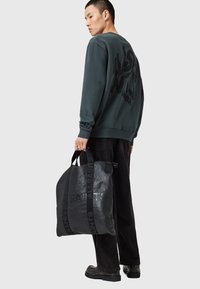 Černá kožená taška přes rameno s embosovaným logem "ALLSAINTS", pevnými uchy a texturovaným povrchem, kterou drží osoba oblečená v zelené mikině.