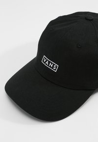 Cappellino da baseball in cotone nero con visiera curva, dotato di un logo rettangolare bianco con la scritta "VANS". Tessuto texturizzato e fori di ventilazione visibili.