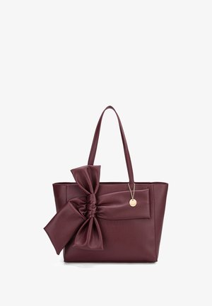 Borsa a tracolla in pelle bordeaux con un grande dettaglio a fiocco sul davanti, fondo piatto e manici lunghi. Charm in tono oro attaccato che aggiunge un elemento decorativo.