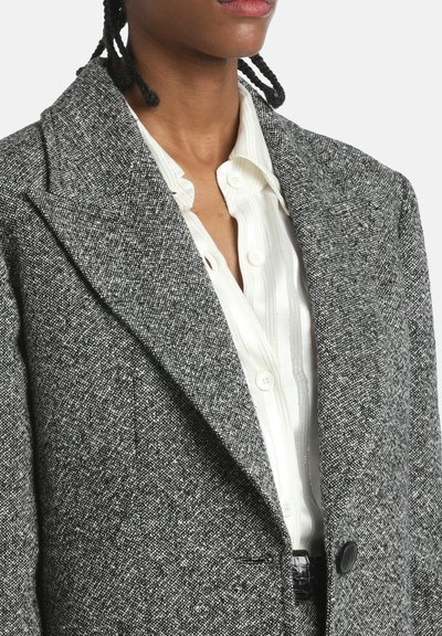 Šedý tvídový blazer s klopovým límcem, jedním knoflíkem a postranními kapsami, nošený přes bílou košili s proužky.
