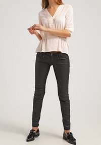 Jasnoróżowa bluzka z krótkim rękawem i plisowanymi detalami, zestawiona z dopasowanymi czarnymi dżinsami. Model nosi czarne mokasyny. Gładki materiał, luźny krój.