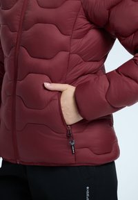 Czerwona pikowana kurtka z falistym wzorem, wyposażona w zamek błyskawiczny z przodu, kieszeń na ręce oraz elastyczne mankiety. Gładkie, błyszczące wykończenie.