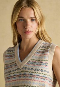 Blonde Frau mit blauen Augen, die einen beigen, gestrickten ärmellosen Pullunder mit mehrfarbigen Streifen und Mustern vor einem schlichten Hintergrund trägt.