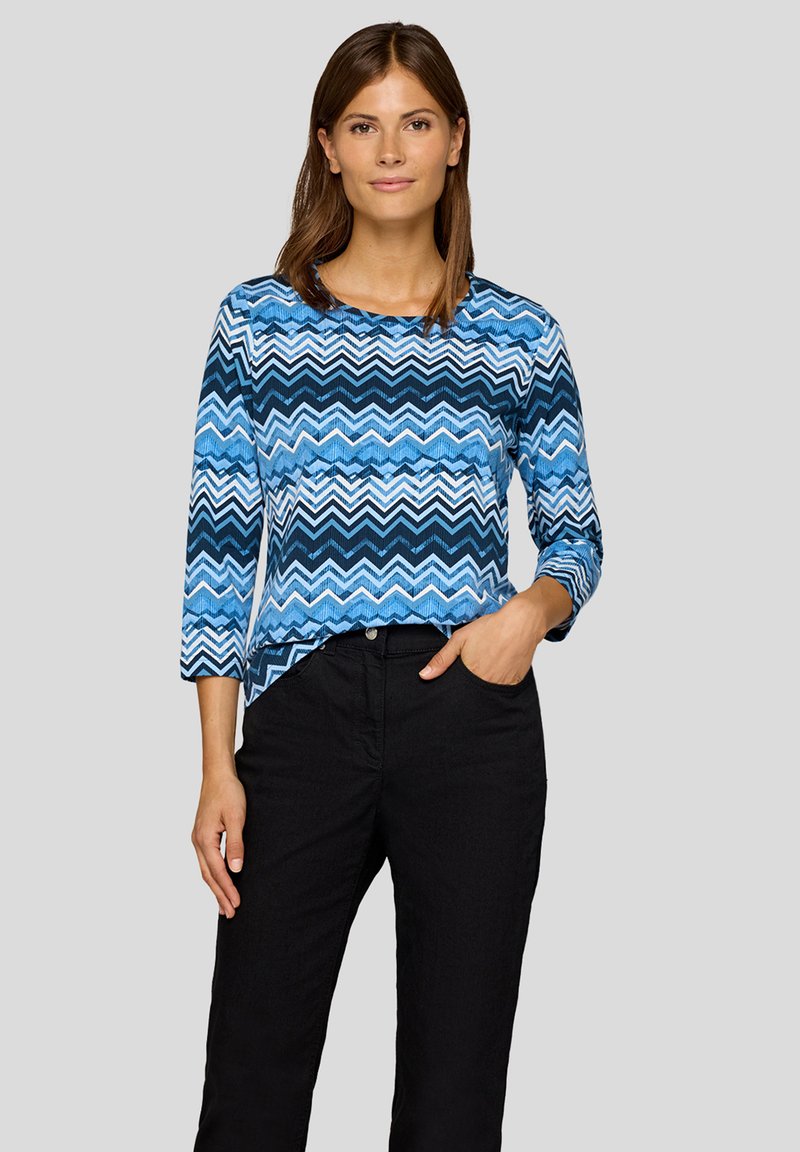 Bluse mit zigzagförmigem Muster in Blau, mit Dreiviertelärmeln und rundem Ausschnitt, kombiniert mit schwarzen Hosen. Weiches Gewebe.