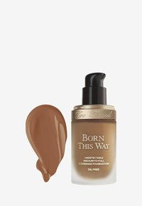 Too Faced Born This Way foundation in een heldere glazen fles met een zwarte pomp, voorzien van gouden accenten. Een swatch van mediumbruine foundation ernaast.