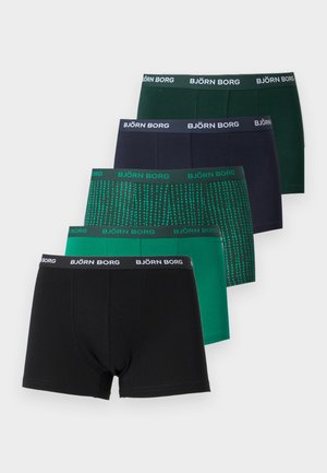 Homme athlétique debout pieds nus sur un sol moquetté, portant un boxer noir Björn Borg, regardant sur le côté avec une main sur la hanche.