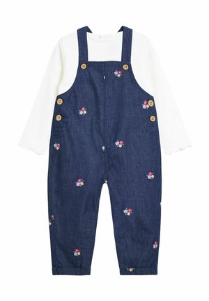 Weißes Langarmhemd, kombiniert mit navyblauen Jeanslatzhosen, die mit kleinen Blumenstickereien und braunen Knöpfen an den Trägern und Seiten verziert sind.