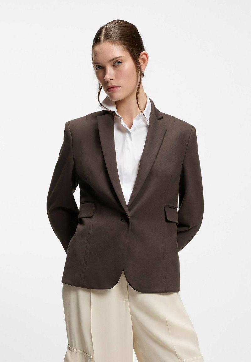 Femme portant un blazer marron par-dessus une chemise blanche et un pantalon large crème, debout les mains derrière le dos sur un fond uni.