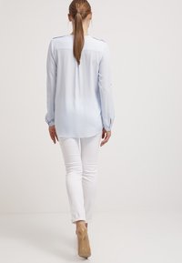 Ljusblå långärmad blus med rundad fåll och knäppta ärmslut, matchad med figurnära vita byxor och transparenta högklackade skor.