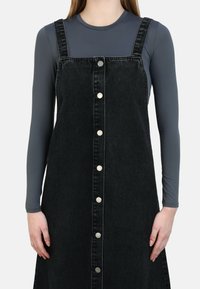 Abito salopette in denim nero con bottoni frontali argentati, bretelle regolabili e una vestibilità ampia a trapezio. Indossato sopra una maglia a maniche lunghe grigia.