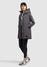 Femme portant un manteau à capuche gris, des leggings noirs, des baskets blanches et noires, et une casquette de baseball noire, debout les mains dans les poches.