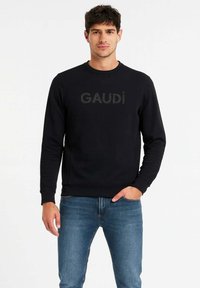 Felpa nera a girocollo con logo "GAUDI" in una texture leggermente in rilievo, abbinata a jeans blu chiaro. Tessuto liscio, vestibilità rilassata.