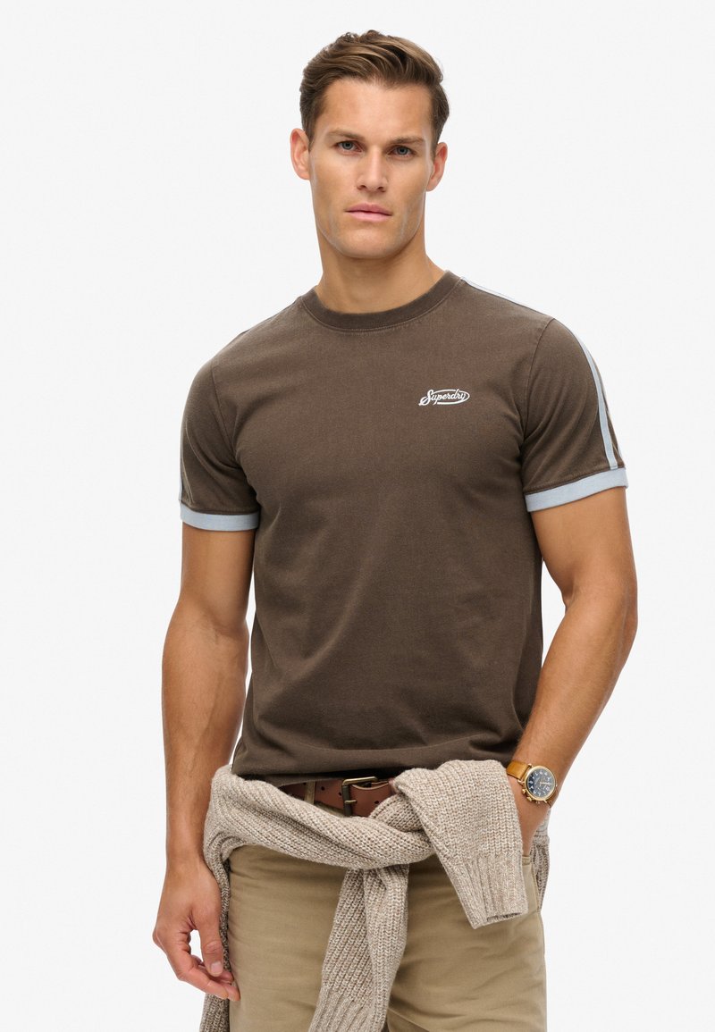 Camiseta de algodón marrón con mangas acanaladas azul claro y un logo blanco en el pecho. Combinada con pantalones caqui y un suéter beige atado a la cintura.