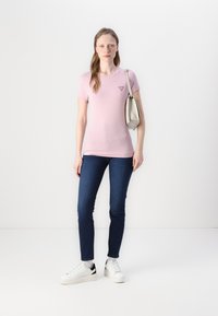 T-shirt rosa chiaro a maniche corte con un piccolo logo triangolare, abbinato a jeans blu scuri e sneakers bianche con dettagli neri.