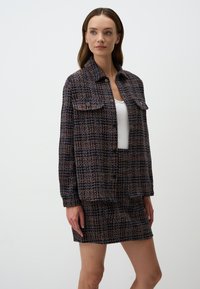 Veste en tweed texturé dans des teintes marine, orange et crème, dotée de deux poches frontales et de fermetures à pression. Associée à une mini-jupe assortie.
