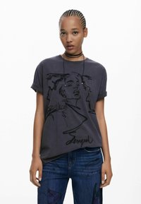 T-shirt en coton gris foncé avec des manches retroussées, comportant un dessin de visage en ligne noire et le texte "Desigual" au bas.