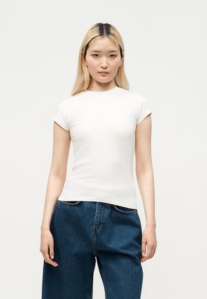 Ung kvinde med langt lyst hår iført en tætsiddende hvid kortærmet skjorte og løstsiddende mørkeblå jeans, der står foran en hvid baggrund.