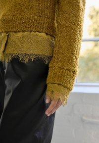 Gestrickter senffarbener Pullover mit strukturierter Oberfläche über einer blütenspitzengesäumten Bluse, schwarze Lederhose mit detaillierten Spitzenkanten an den Ärmeln.