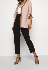 Vaaleanpunainen kimono-tyylinen takki, jossa on paljettikoristereuna, pukeutuneena valkoisen satiinitopin päälle, yhdistettynä mustiin räätälöityihin housuihin ja mustiin hihnakenkiin.