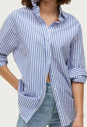 Persona vestida con una camisa de botones azul y blanca a rayas verticales, con las mangas arremangadas, y jeans de un azul claro, con la mano descansando en la cadera.