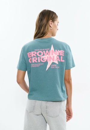 Femme aux cheveux bruns portant un T-shirt bleu canard avec un grand texte rose « Brownie Original » et un motif étoile, associé à un jean gris clair, vue de dos.