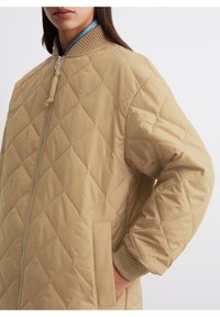 Kobieta w beżowej, pikowanej kurtce z ribbowanym kołnierzem i mankietami, z zamkiem z przodu, trzymająca ręce w kieszeniach bocznych, pokazana od szyi do bioder.