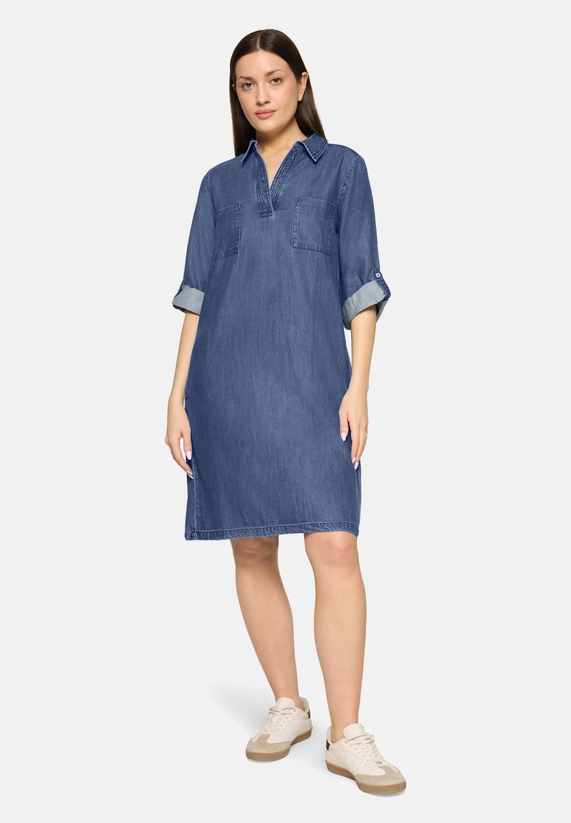 Femme portant une robe en denim bleue longueur genou avec des manches retroussées et des baskets blanches, debout devant un fond clair uni.