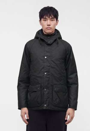 Young man wearing a black hooded jacket with snap buttons and front flap pockets, standing against a plain light background.