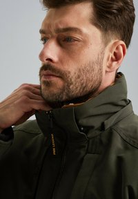 Olijfgroene jas met een hoge kraag, ritssluiting en een gele ritstrekker. De stof heeft een subtiele structuur en is ontworpen voor warmte en stijl.