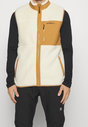 Man met een wit- en beige fleecevest met borstzak met rits, een zwart shirt met lange mouwen en een zwarte broek, tegen een effen achtergrond.